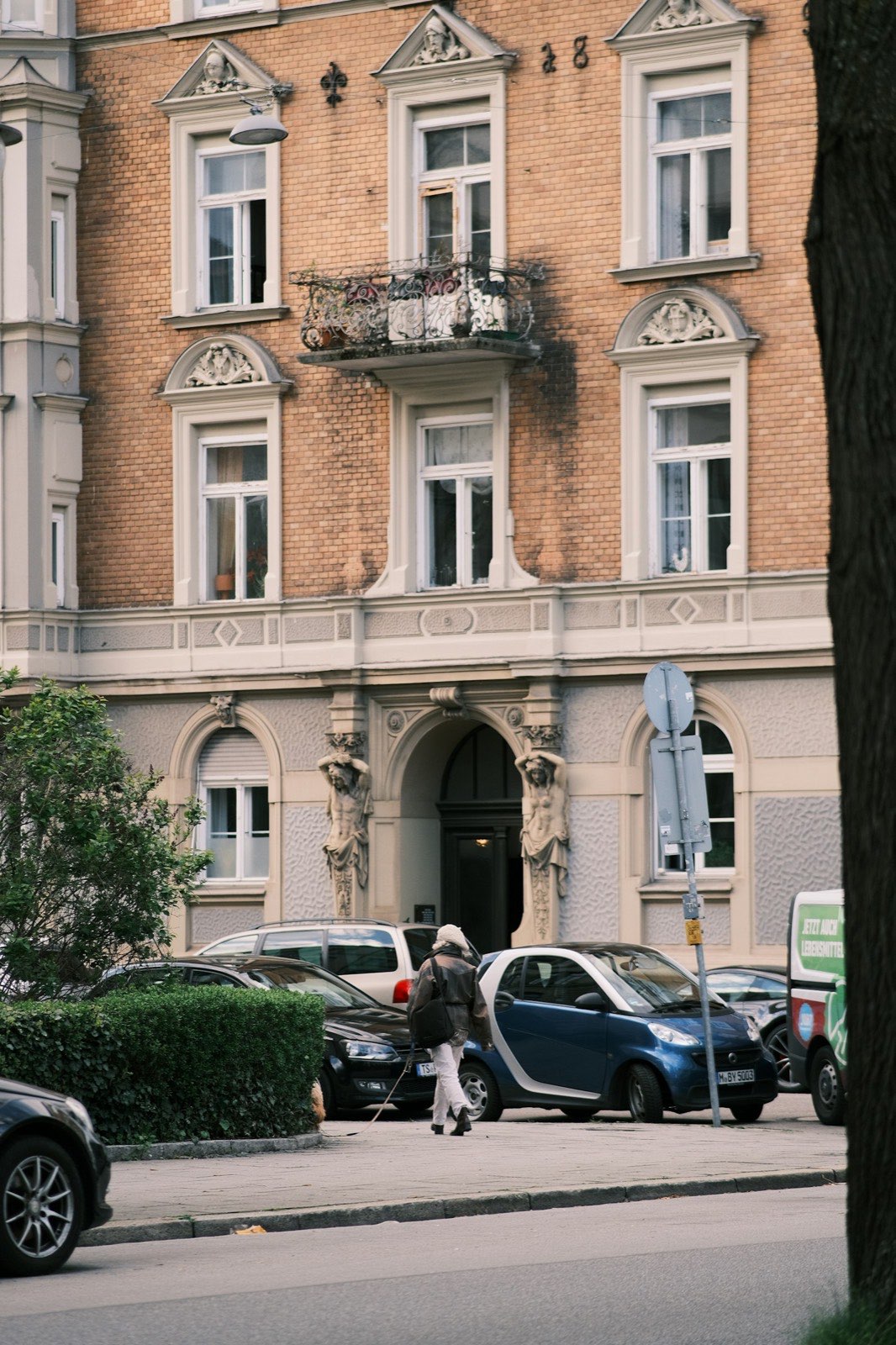 Münchner Altbau an einer Wohnstraße, davor parkende Autos und eine Person auf dem Gehweg